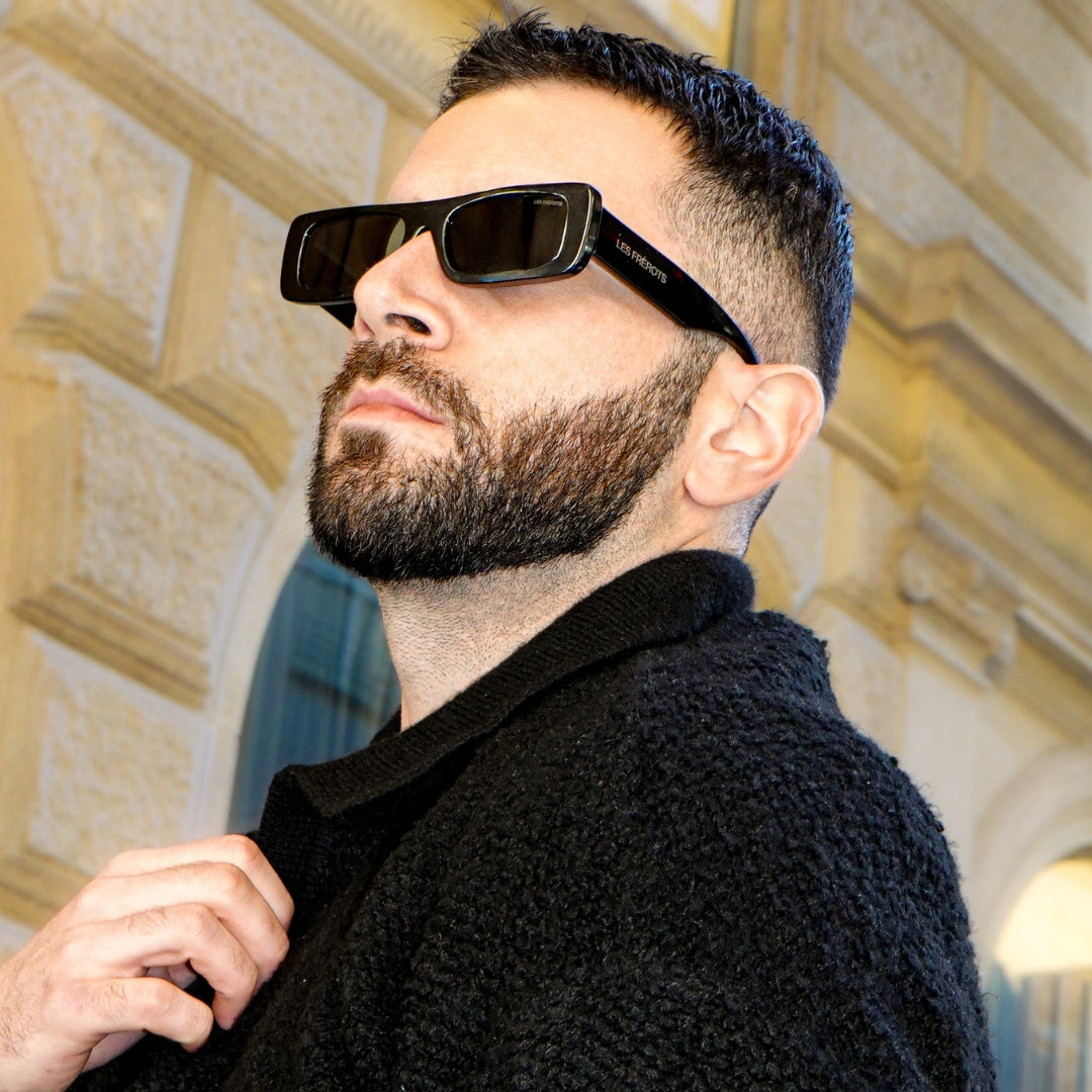 Man wearing black sunglasses and a dark coat in front of an architectural background